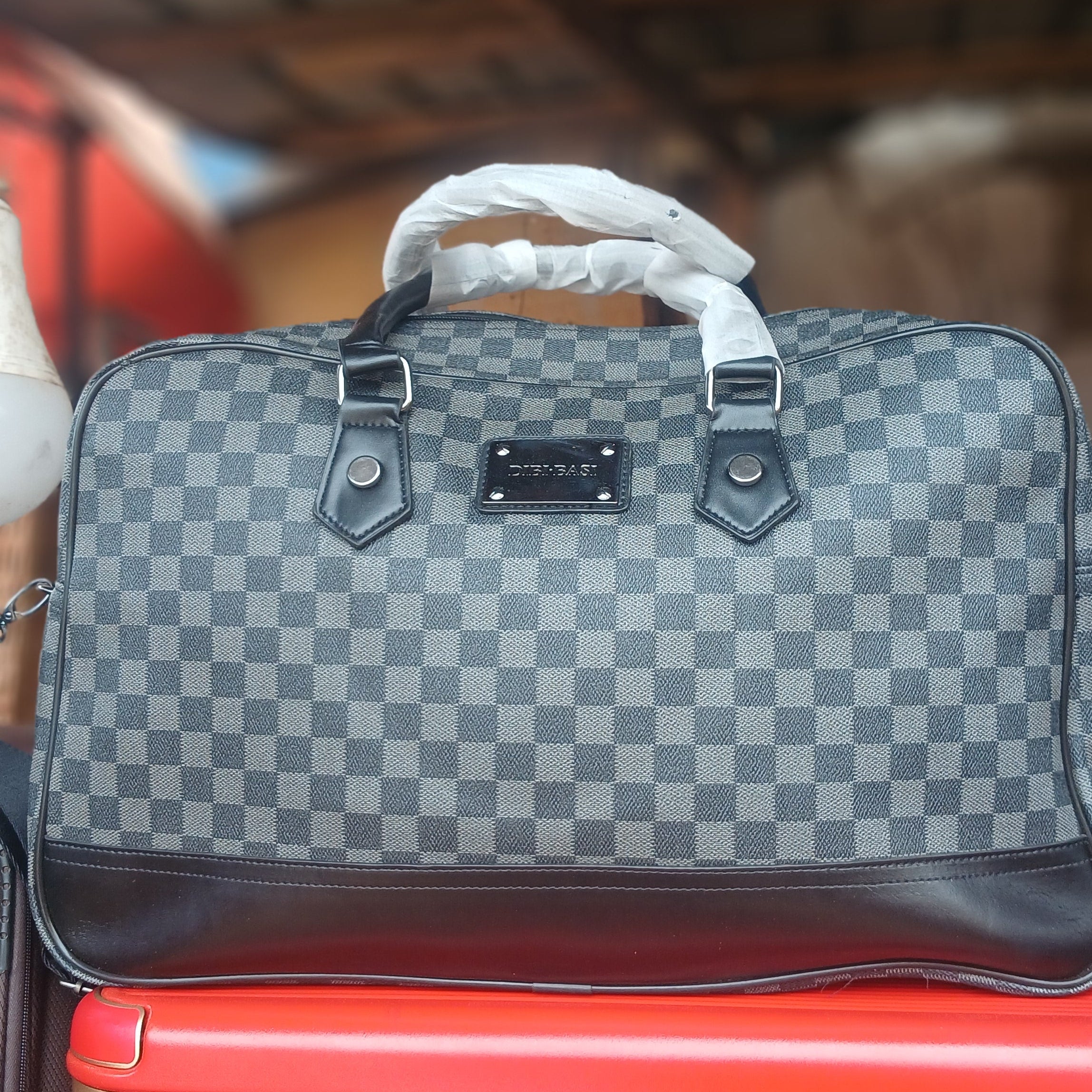 gray and black checkerboard pattern travel holdall bag featuring dark leather trim and a central logo plaque.